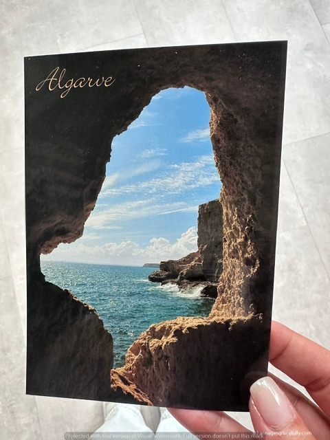Een ansichtkaart van de Algarve met een uitzicht op de oceaan door een natuurlijke opening in de rotsen, onder een heldere blauwe lucht.