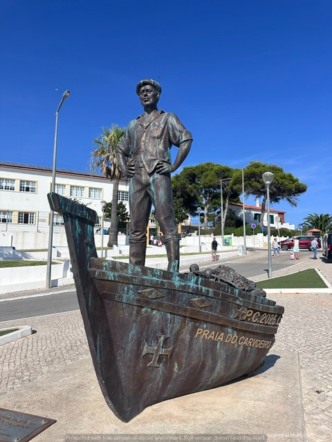 Bronzen standbeeld van een man in maritieme kleding, staande op de boeg van een schip, met een blauwe lucht en palmbomen op de achtergrond, gelegen bij Praia do Carvoeiro.