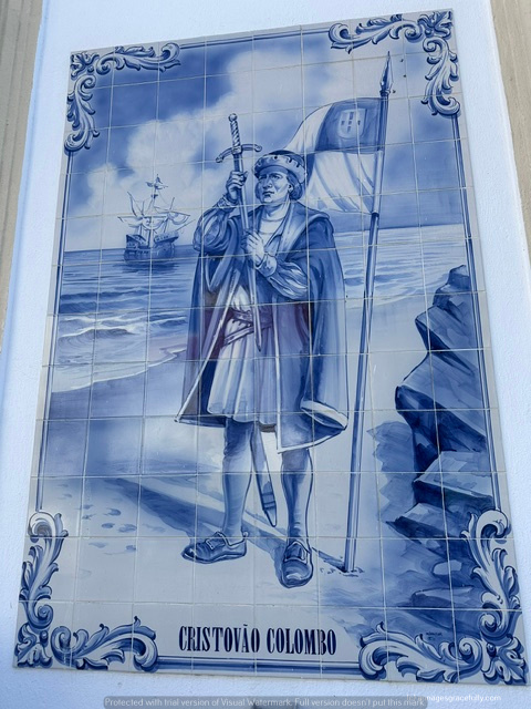 Een tegeltableau van Christoffel Columbus, staande aan de kust met een zwaard en een vlag, met een schip op de achtergrond.