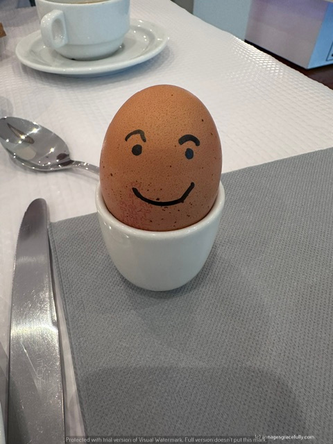 Een gekookt ei met een lachend gezichtje, geplaatst in een wit eitjehouder, op een ontbijttafel met bestek en een lege kop koffie op de achtergrond.