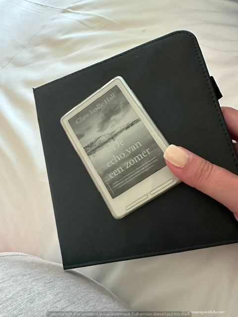 Een hand houdt een e-reader met de titel 'De echo van een zomer' van Clare Ludlike Hall, gelegen op een bed met een neutrale achtergrond.