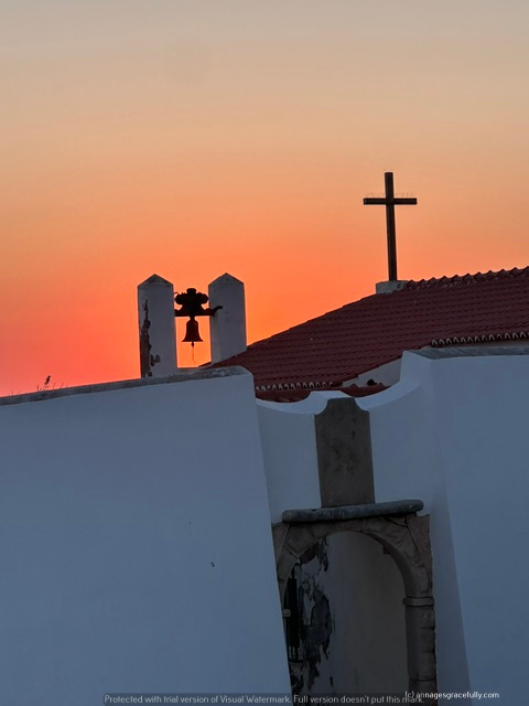 Een silhouet van een kerk met een klokkentoren en een kruis tegen een kleurrijke zonsondergang.
