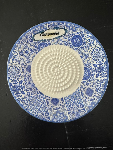 Een ronde blauwe en witte schotel met een gedetailleerd, geometrisch patroon en een centrale spiraaltextuur. De schotel heeft een etiket met de naam 'Carvoeiro'.