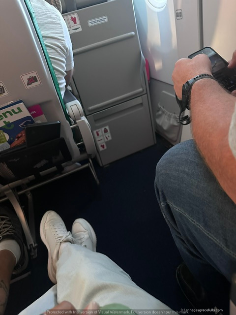 Zicht op de benen van passagiers in een vliegtuig, met de focus op witte sneakers en een arm die een smartphone vasthoudt.