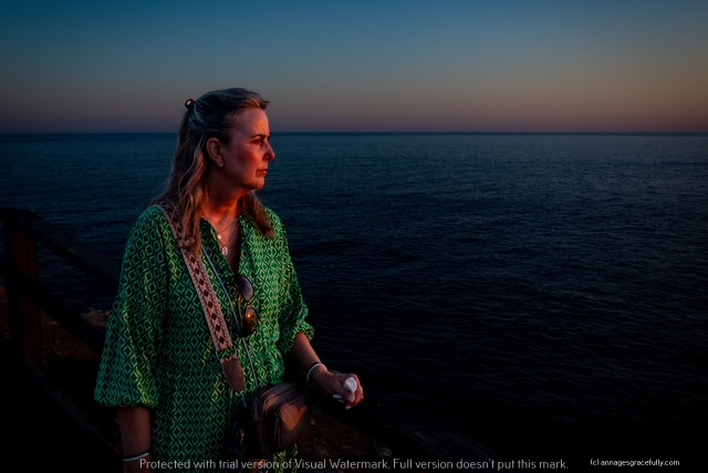 Een vrouw met halflang haar in een groene blouse staat naast een hek aan de kust, kijkend naar de zee tijdens de schemering.