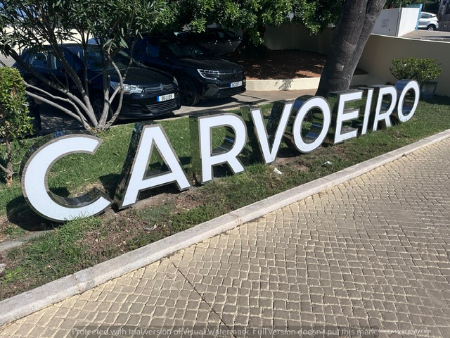 Een grote, witte sign met het woord 'CARVOEIRO' in hoofdletters, geplaatst op een groene grasstrook naast een parkeerplaats.