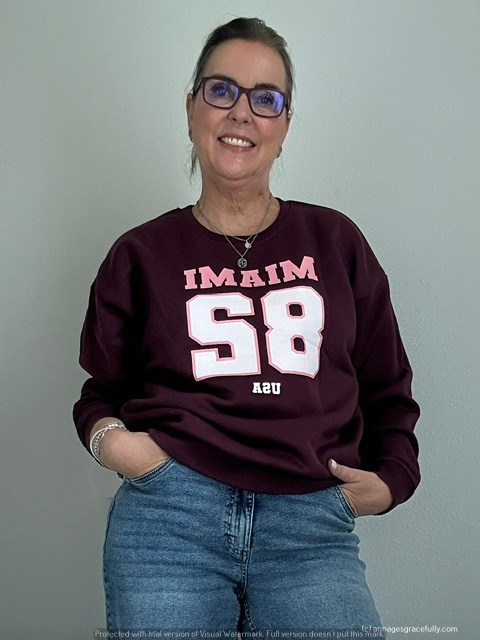 Een glimlachende vrouw met een korte, stijlvolle hairstyle, draagt een donkerbruine sweater met de tekst 'IMAI 58' en een spijkerbroek. Ze heeft een moderne bril op en poseert met een ontspannen houding tegen een lichte achtergrond.