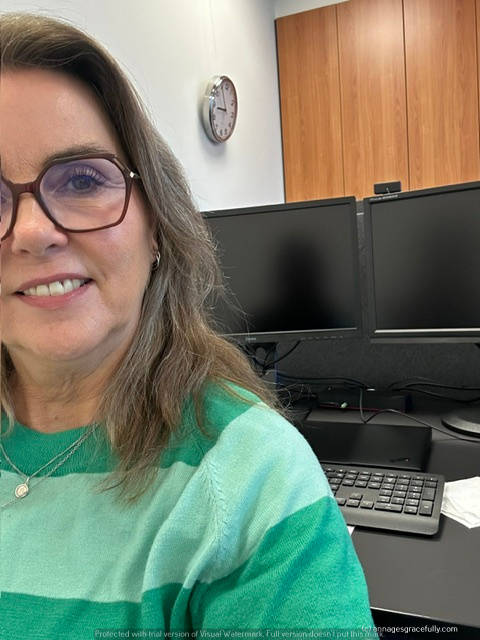 Een vrouw met een groen gestreepte trui, die glimlacht terwijl ze voor een bureau met twee computerschermen zit. Op de achtergrond hangt een klok op de muur.