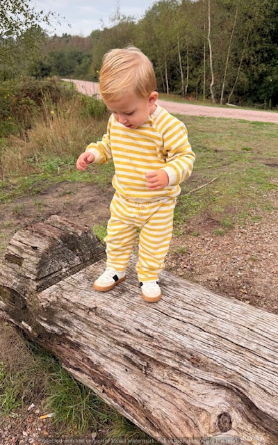 Een peuter in een gestreept geel en wit outfit staat op een boomstam in een natuurlijke omgeving.