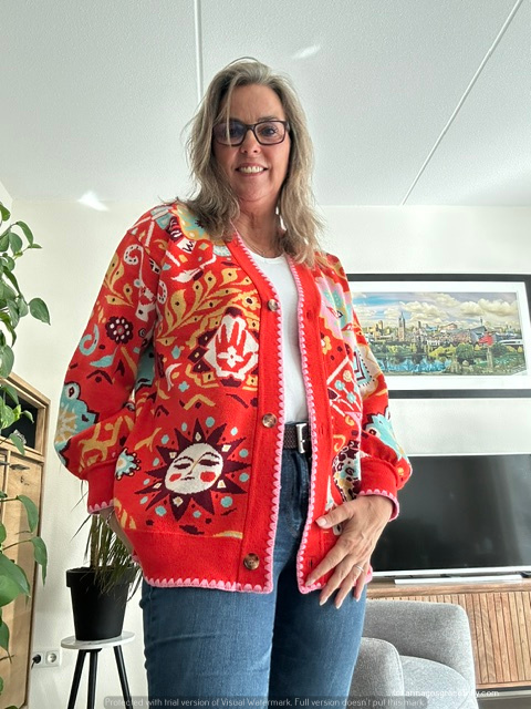 Een vrouw met grijs haar en een bril poseert in een kleurrijke gebreide cardigan met een uniek ontwerp, in een heldere woonkamer.