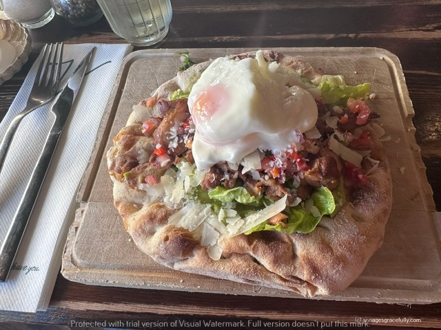 Een plattebrood met toppings zoals gesneden vlees, groenten, kaas en een gebakken ei, geserveerd op een houten plank.
