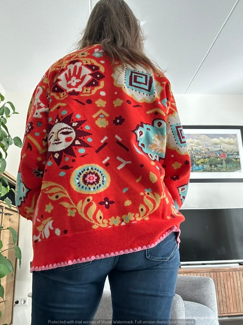 Een vrouw met lange haren draagt een felrode trui met kleurrijke patronen, terwijl ze met haar rug naar de camera staat.