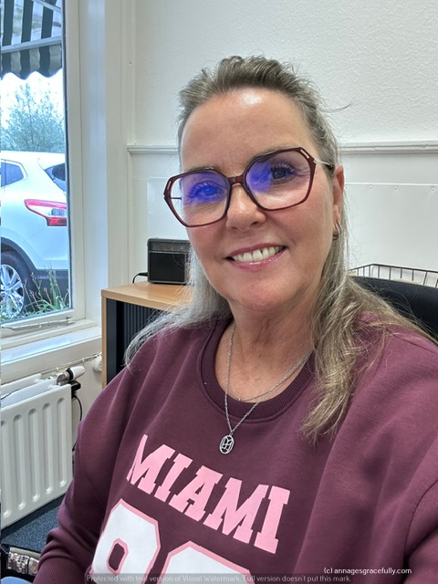 Vrouw met lang haar en bril, zittend aan een bureau met een glimlach, gekleed in een sweatshirt met de tekst 'MIAMI'. Op de achtergrond is een raam met uitzicht op een parkeerplaats.