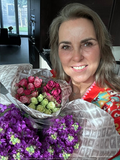Een vrouw poseert vrolijk met een bos bloemen in haar handen, bevat rozen en paarse bloemen, in een moderne keukensetting.