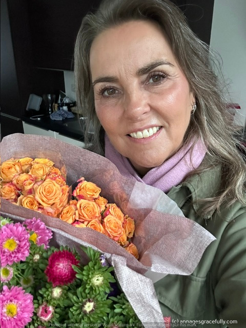 Een vrouw lacht terwijl ze een boeket bloemen vasthoudt, inclusief oranje rozen en kleurrijke asters, met een gezellige achtergrond.