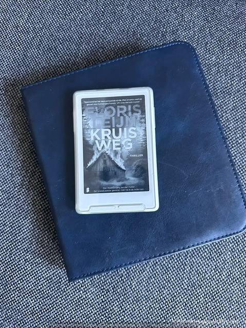 Een e-reader met de cover van het boek 'Kruisweg' van Morris Teijne, gelegd op een blauw leren boek in de buurt van een textuurrijke ondergrond.