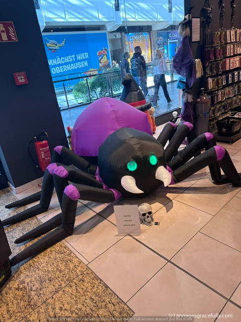 Een opblaasbare spin met paarse en zwarte kleuren, met groene ogen en witte tanden, staat in een winkel, omringd door Halloween-decoraties.