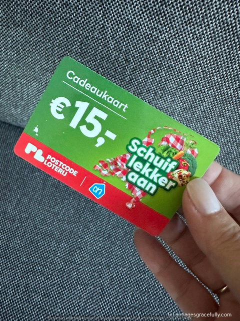 Een hand die een cadeaukaart vasthoudt, met een waarde van €15,- van de Postcode Loterij, met de tekst 'Schuif lekker aan'.