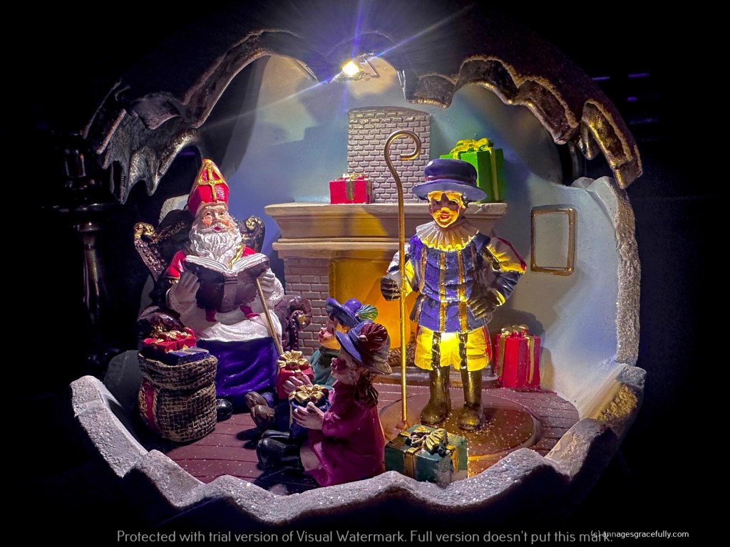 Een kerstdecoratie met Sinterklaas die een boek leest, terwijl Zwarte Piet in een kleurrijk kostuum naast hem staat. Er zijn cadeaus en een haard in de achtergrond.