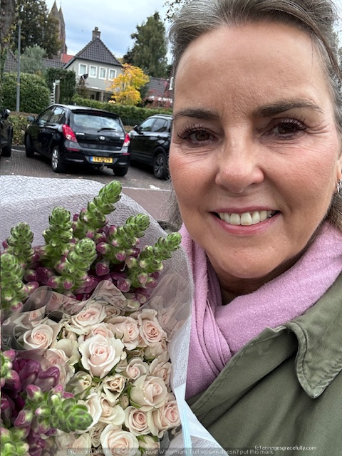 Een vrouw met een glimlach houdt een bos bloemen vast, met roze rozen en groene takken, op een straat met auto's op de achtergrond.