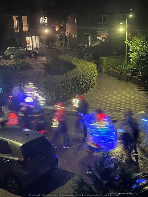 Een avondbeeld van een verlichte straat waar mensen met felgekleurde lichten in beweging zijn, omringd door gebouwen en groene struiken.