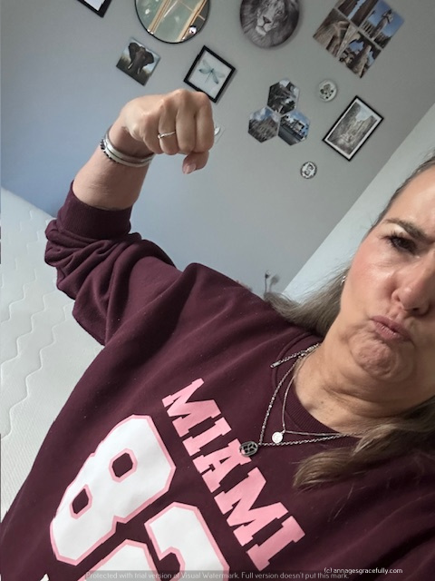 Een vrouw in een bordeaux rood sweatshirt met 'MIAMI 82' erop, die haar arm in een vuistresultaat omhoog houdt, met een krachtige gezichtsuitdrukking, in een kamer met een bed en decoratieve muren.