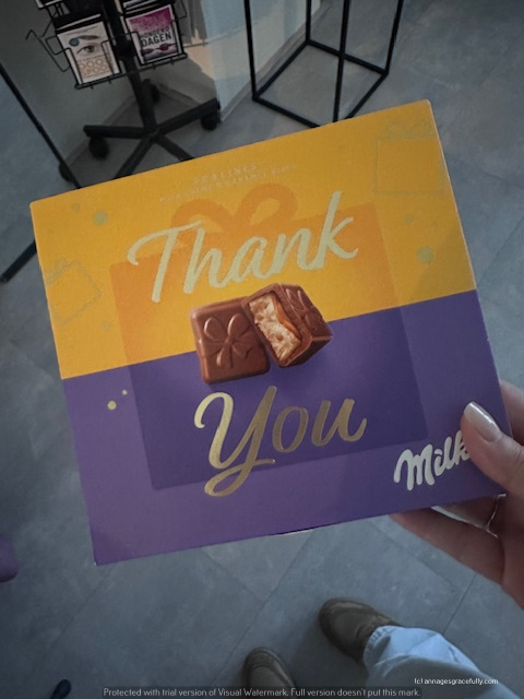 Een hand holdt een geschenkdoos met de tekst 'Thank You' en een praline erop, op een paarse en gele achtergrond.