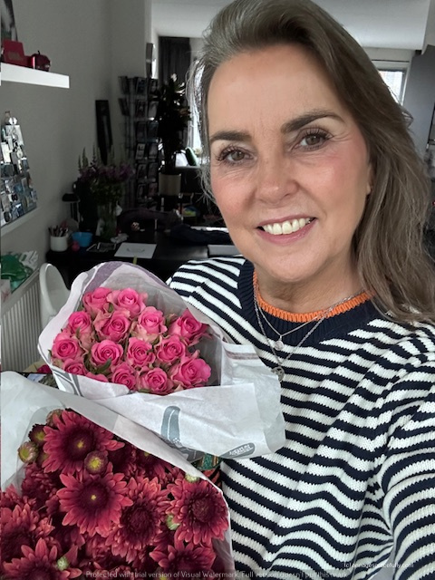 Een vrouw met een glimlach houdt twee boeketten bloemen vast, één met roze rozen en één met paarse chrysanten, terwijl ze zich in een woonkamer bevindt.